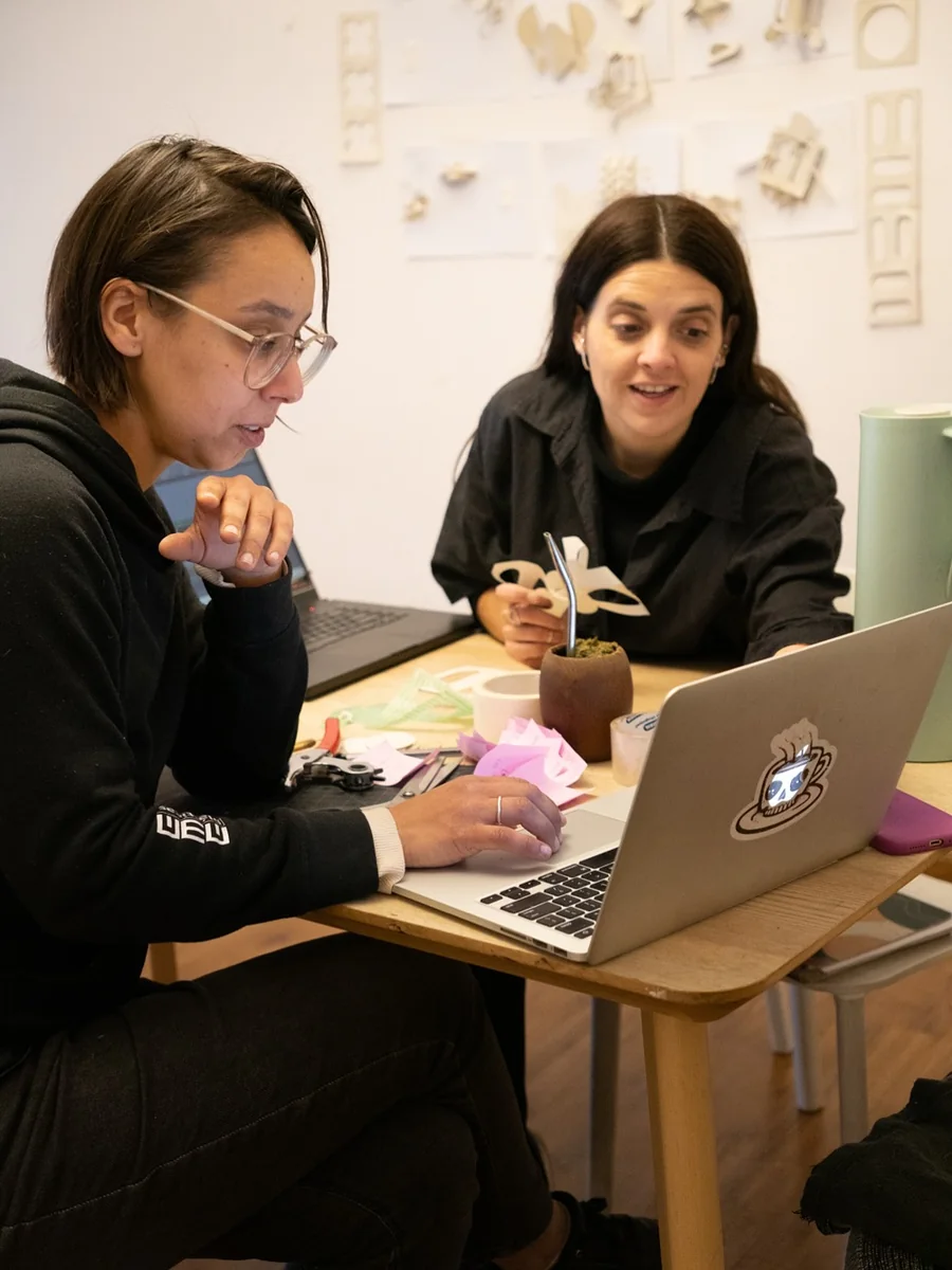 Margarita Martínez y su colaboradora trabajan frente a una computadora, con prototipos de papel sobre la mesa